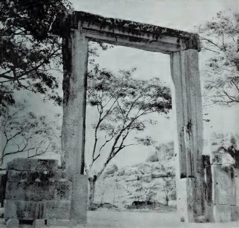 A plain stone gateway of two tall square pillars and a heavy lintel, standing among trees with a rocky hill behind.
