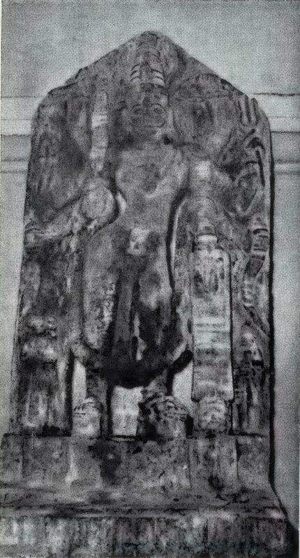 A worn standing god carved on a tall stone slab with a pointed top, with small statues near his feet.