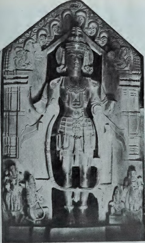 A standing crowned deity under a carved arch on a stone slab with a pointed top, with small attendant statues at the lower corners.