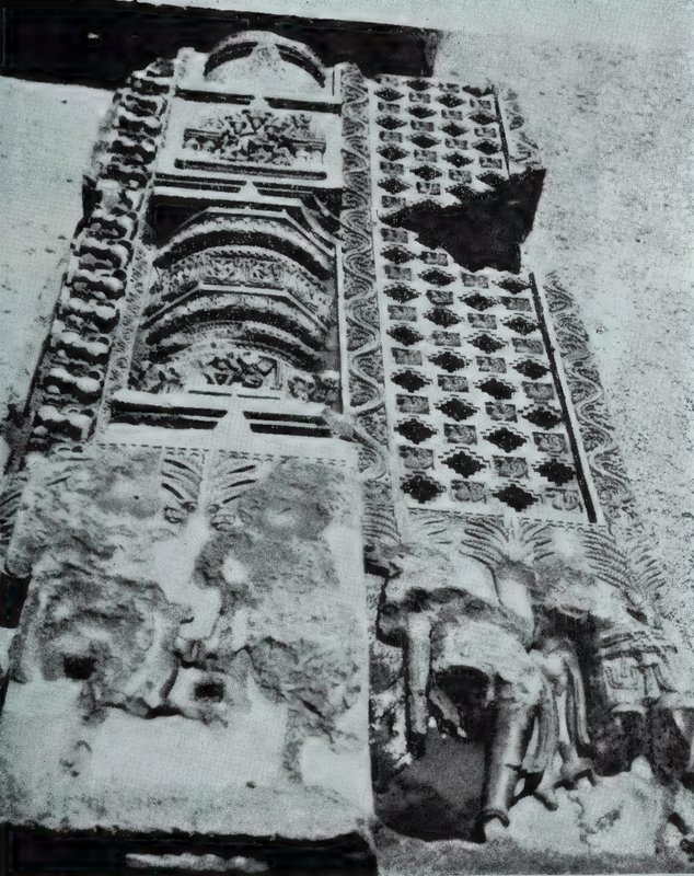 A damaged carved slab seen at an angle, with a pilaster crowned by a small tower motif and a lattice of diamond-shaped openings beside it.