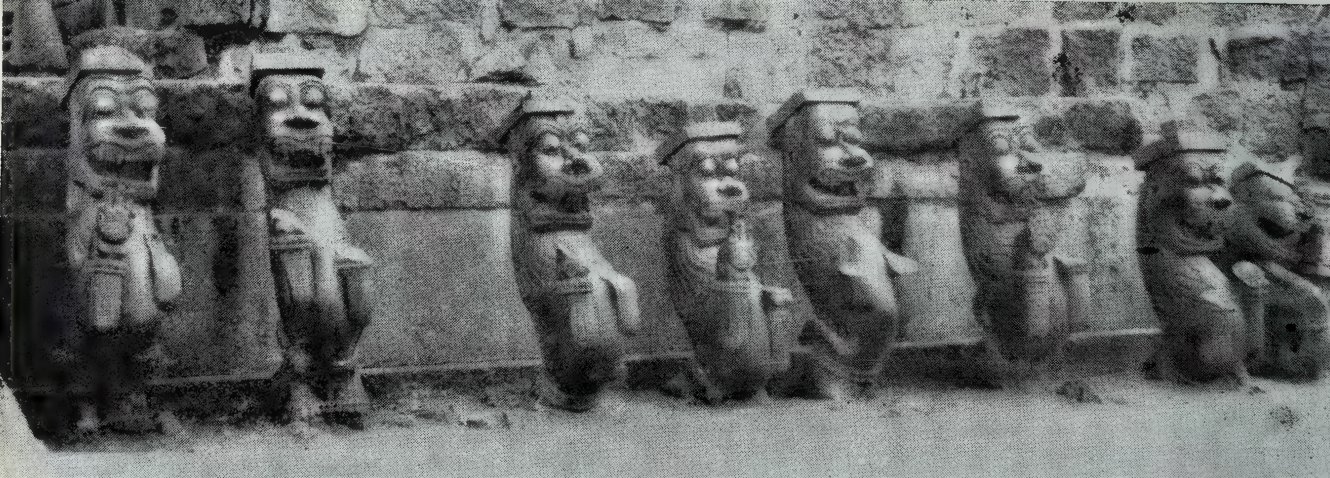 A row of seven weathered stone lion brackets standing in front of a masonry wall.