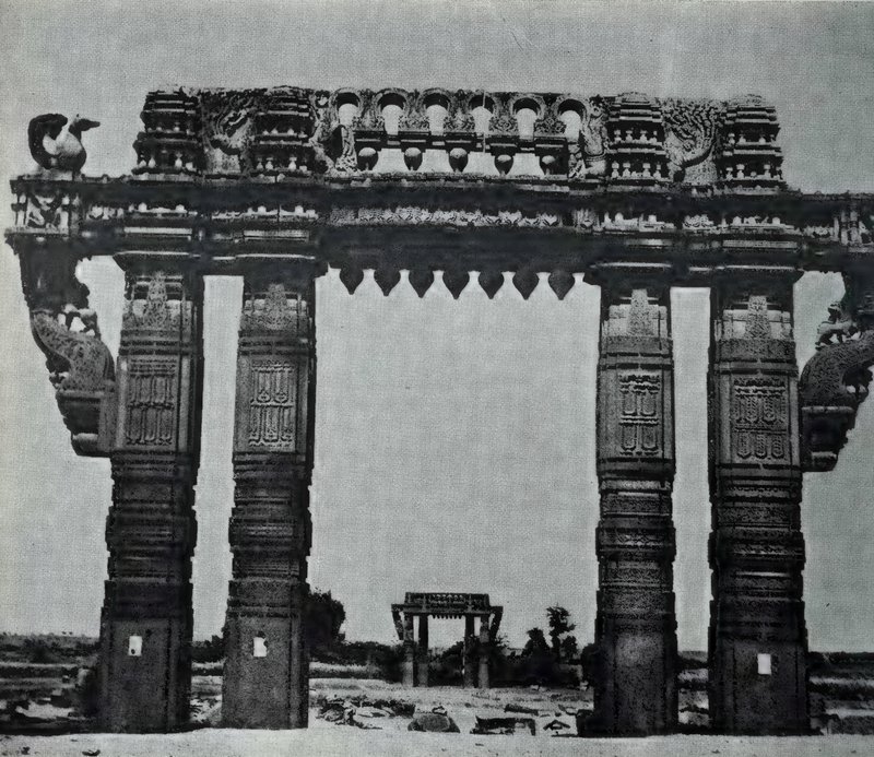A tall stone gateway of four carved pillars carrying a richly carved lintel, with a second gateway visible in the distance.