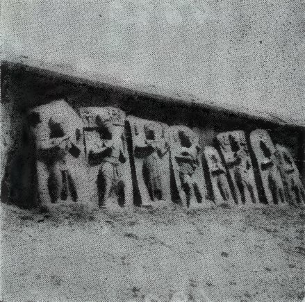 A row of weathered carved slabs with standing people, set at the foot of a low wall.