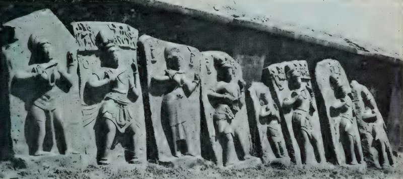 A row of eight carved slabs, each with a standing person, set against a wall.