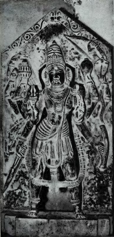 A standing many-armed god holding weapons, carved on a slab with a pointed top, with smaller statues beside him.