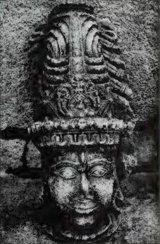 A large carved stone head with wide eyes and a tall headdress of piled, flame-like locks.