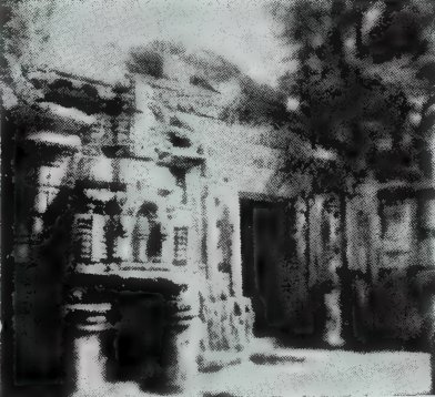 A grainy photograph of a temple front with carved pilasters, a niche and a dark doorway, with trees at the right.
