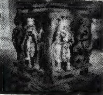 A dark photograph of a square pillar with standing statues carved on its faces.