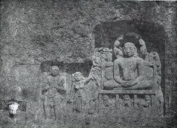 A rock-cut relief on a rough cliff face. A seated person meditates inside a niche, and standing statues are carved to the left.