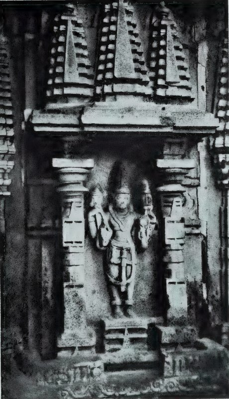 A standing four-armed god in a wall niche between two pillars, below three small carved tower-shaped roofs.