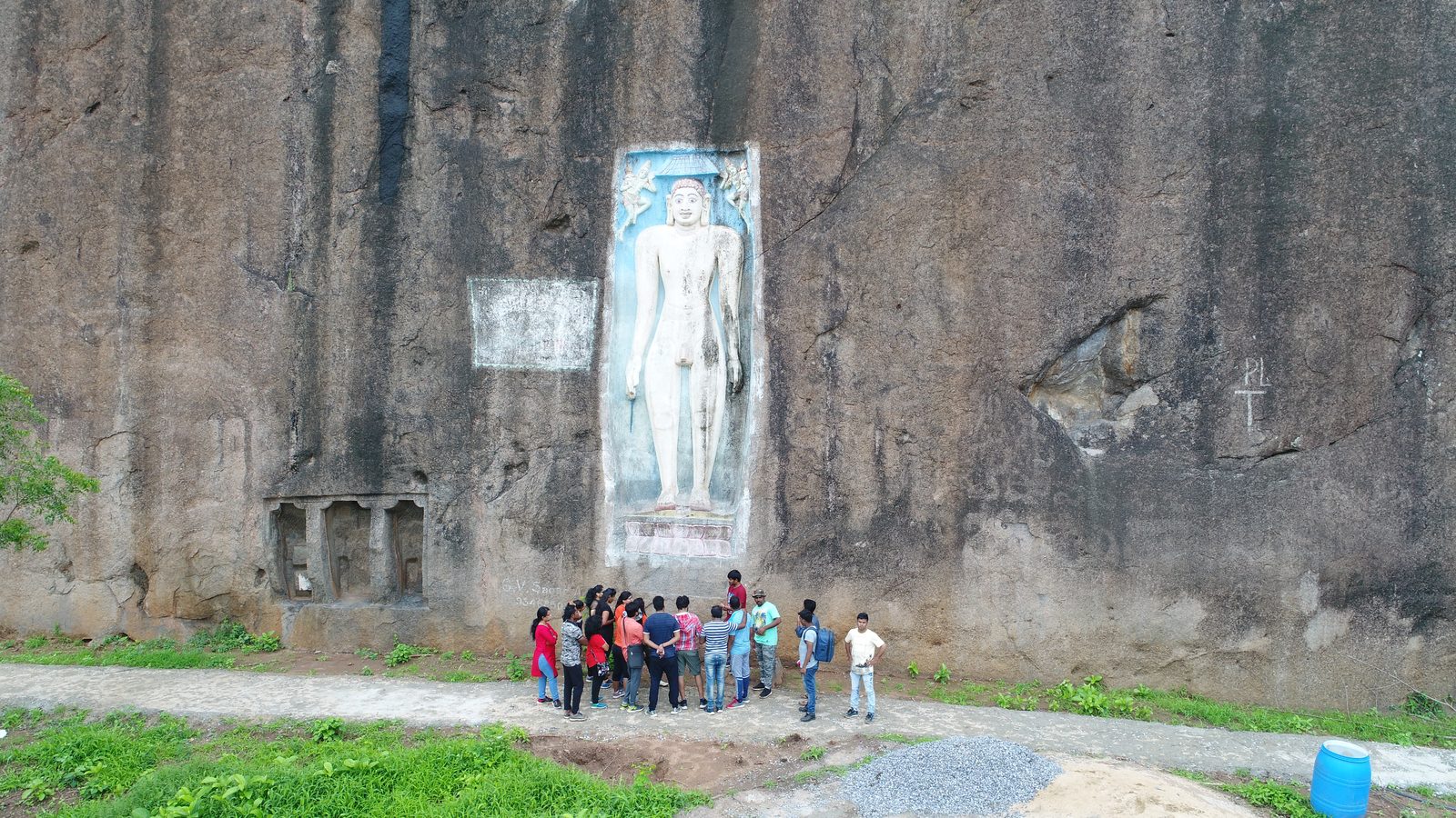 మునిగిపోయిన ఫలకంలో చెక్కబడిన పొడవైన జైన శిల్పంతో కూడిన ముదురు గ్రానైట్ రాతి ముఖం. ఈ విగ్రహానికి నీలం నేలపై తెల్లటి రంగు వేయబడింది. సుమారు పదిహేను మంది సందర్శకుల బృందం దాని పాదాల వద్ద ఒక మార్గంలో నిలబడి ఉంది. దిగువ ఎడమ వైపున ఉన్న రాతిలో చదరపు స్తంభాలతో కూడిన చిన్న ద్వారం కత్తిరించబడుతుంది.