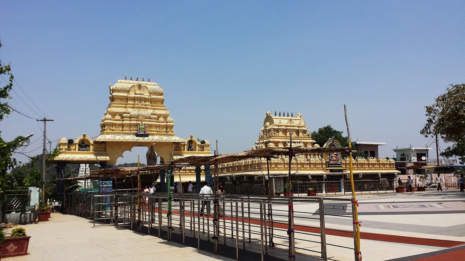 Two cream-and-gold tiered gateway towers rise above a long, low pillared hall. A paved forecourt with metal queue railings lies in front, under a clear blue sky.