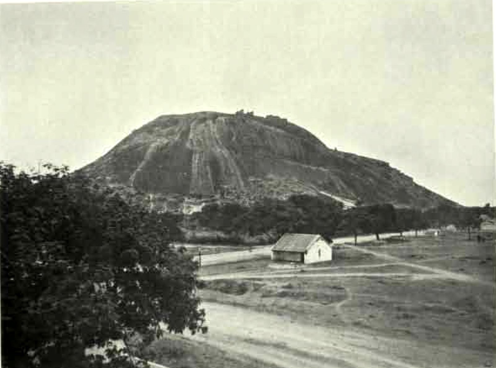 A large rounded rock hill rises behind open ground. A small building with a pitched roof stands beside a road. A tree fills the left foreground.
