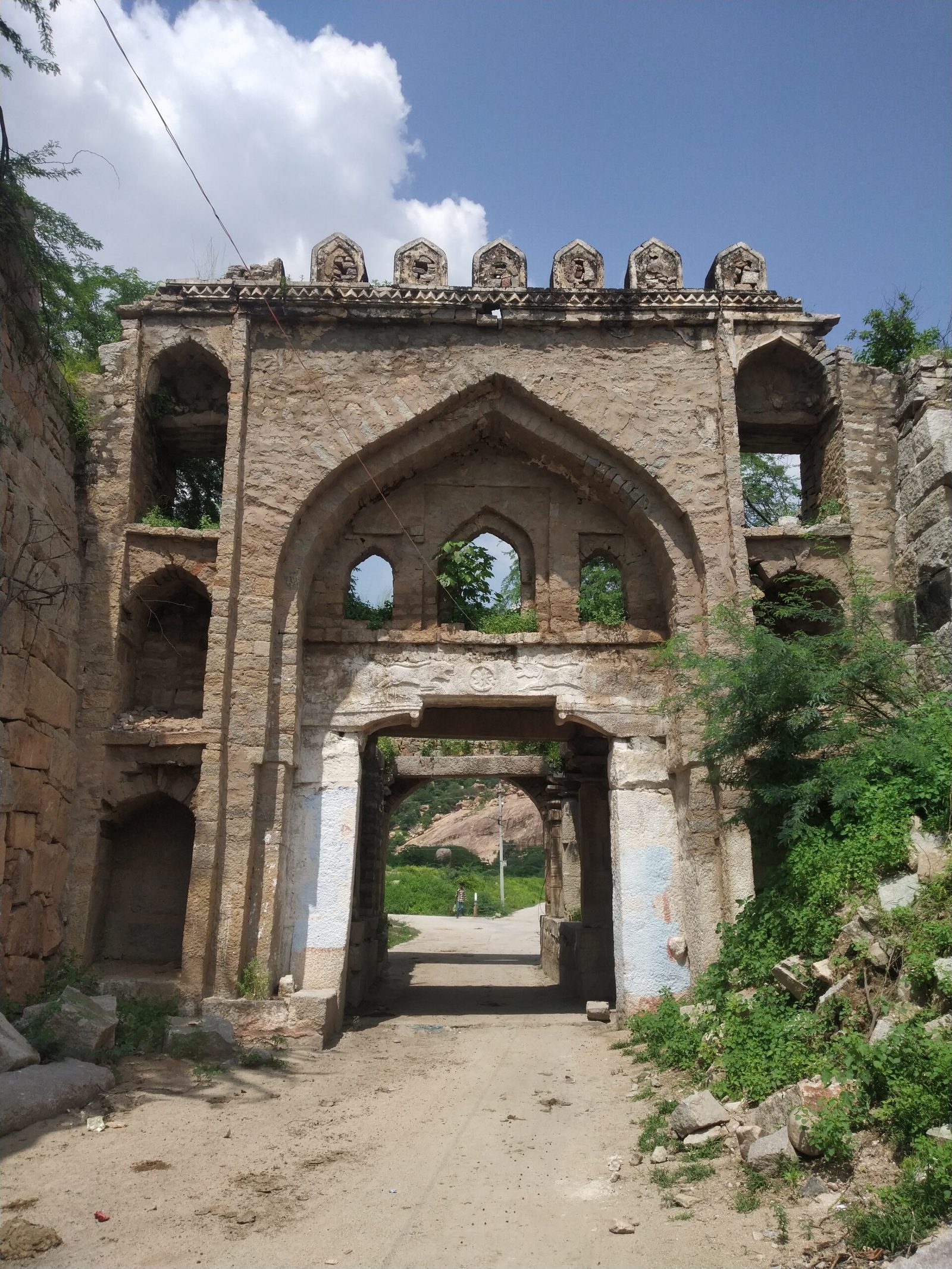 A tall stone gateway has a large pointed arch above a smaller square doorway. Three small arched openings sit inside the large arch. A row of carved stone blocks runs along the top edge, and a dirt road passes through the doorway.