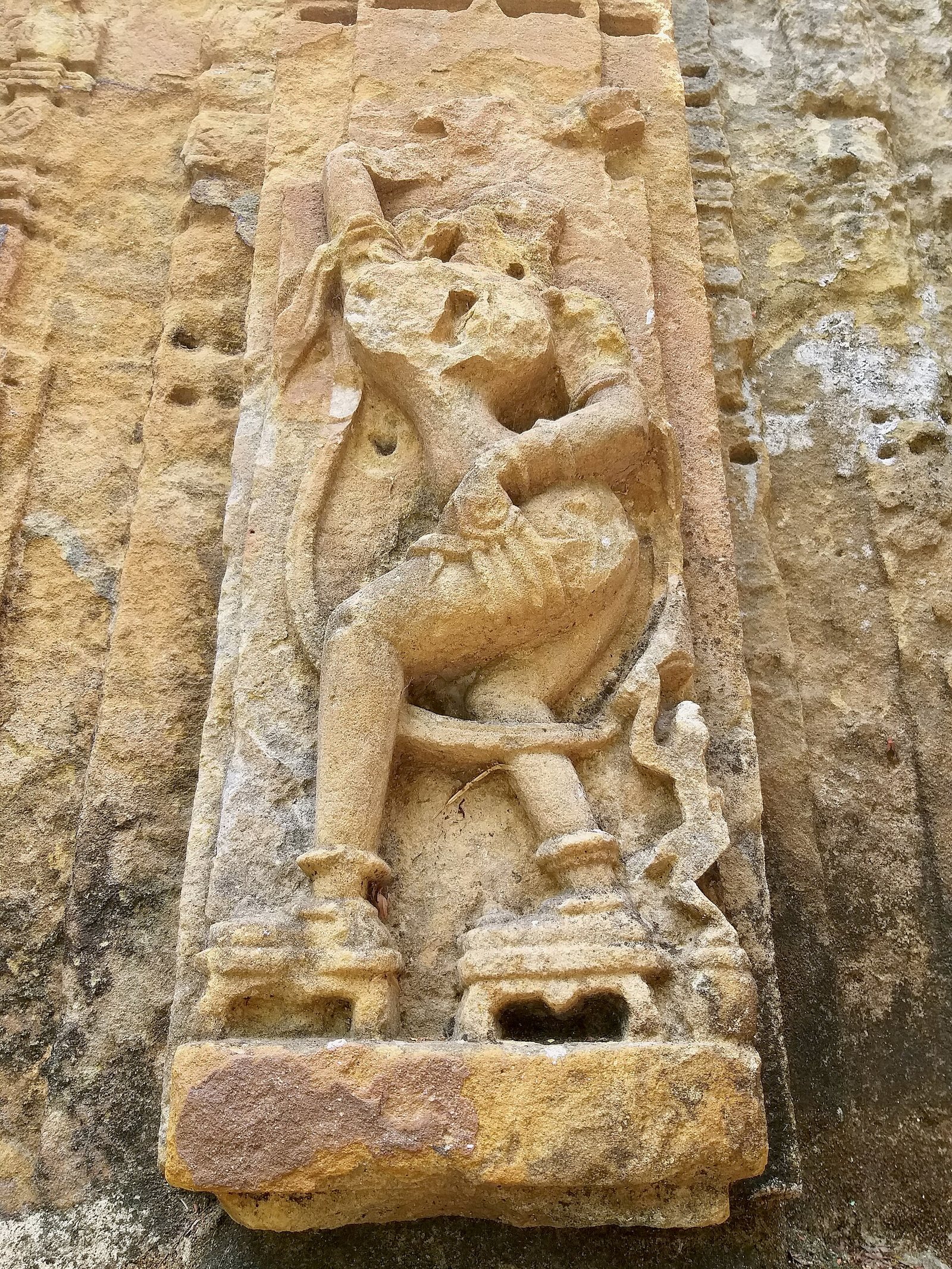 A weathered sandstone panel with a slender sculpture standing in a dancing pose inside a narrow frame.