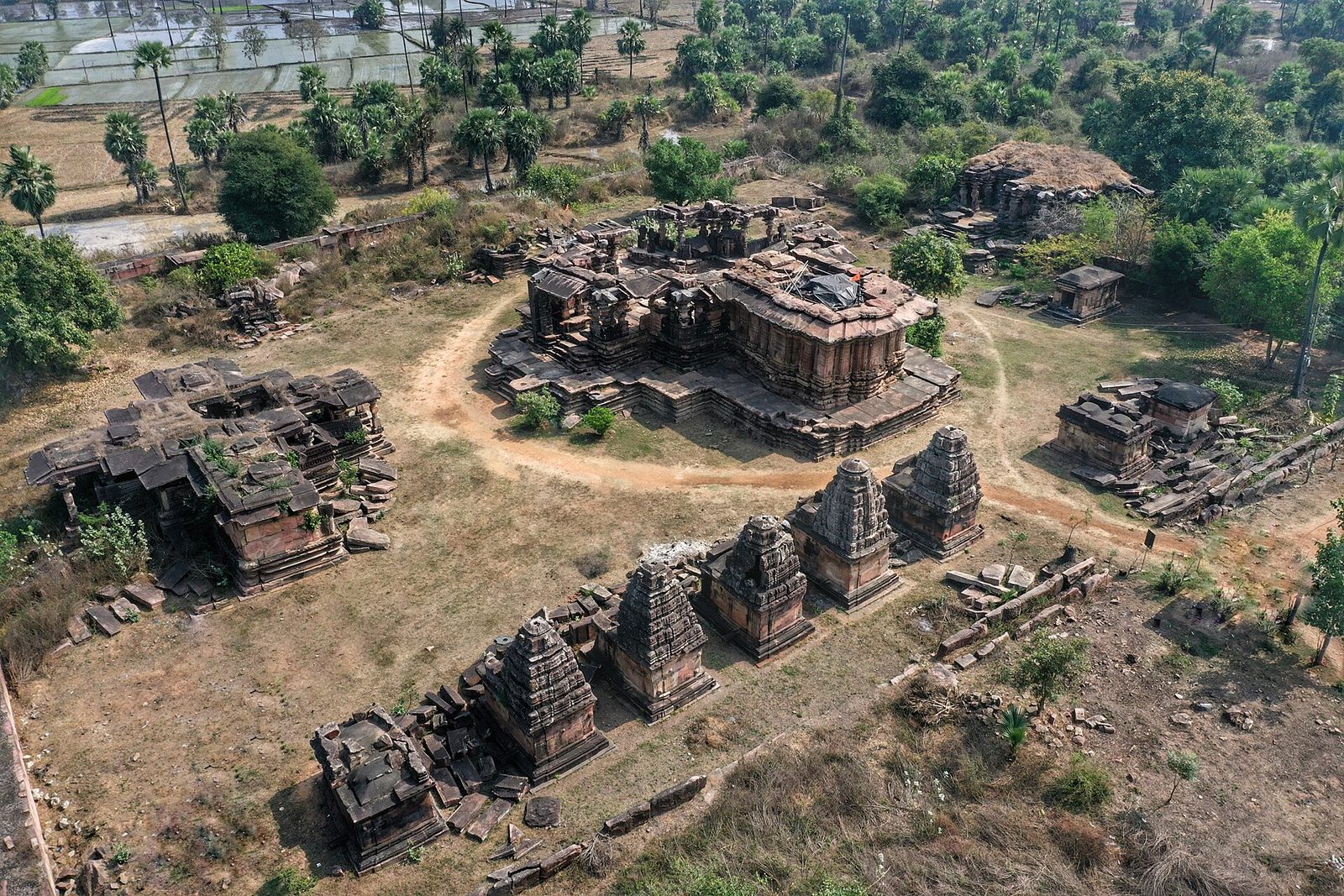 Aerial view of the Ghanpur temple complex: a central temple on a large platform, smaller shrines with towers around it, ruined structures and paths, set among fields and palms