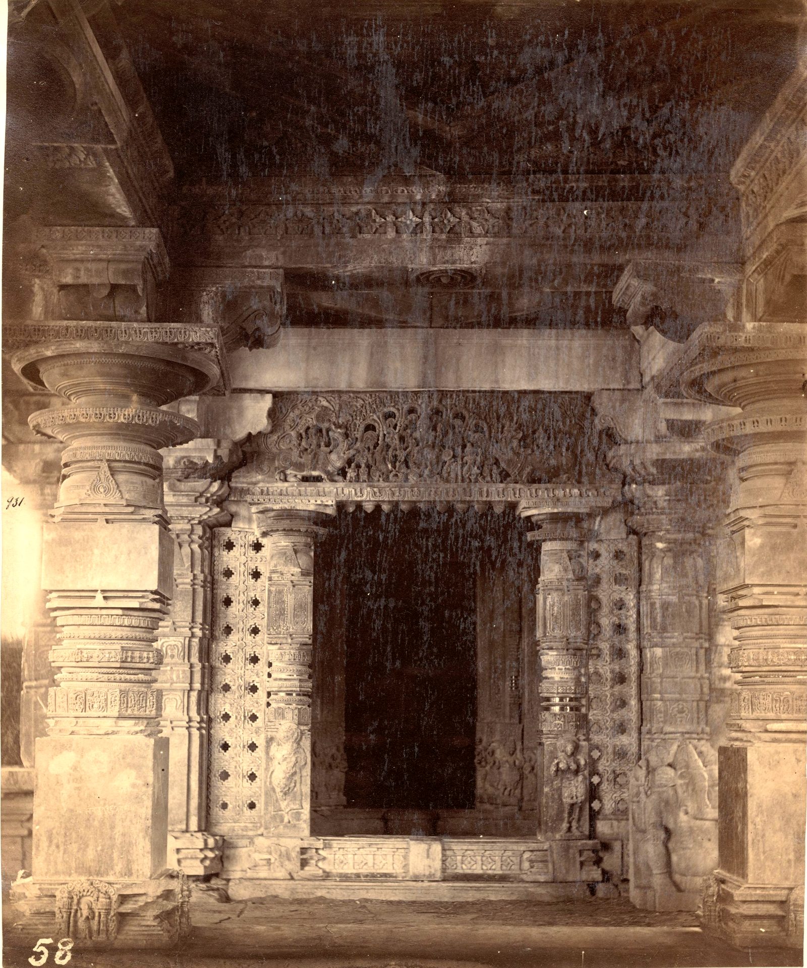 A dark shrine doorway framed by carved jambs and pierced stone screens. Heavy carved pillars and a carved lintel surround the doorway.