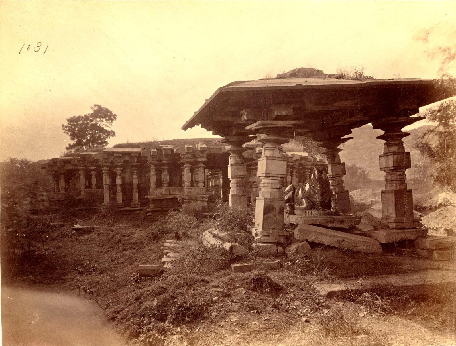 A stone pavilion with a heavy flat roof stands on carved pillars on uneven ground. A long pillared building stands behind it at the left. Rubble and loose stones lie in front.