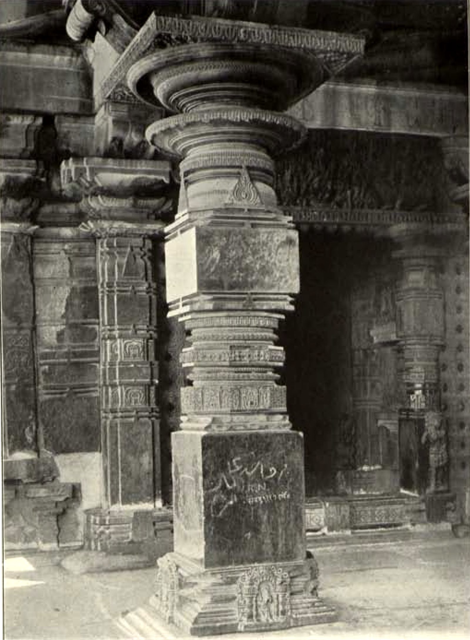 A tall carved stone pillar with a wide round capital and a square base inside the temple. Writing in two scripts is scratched on the base of the pillar.