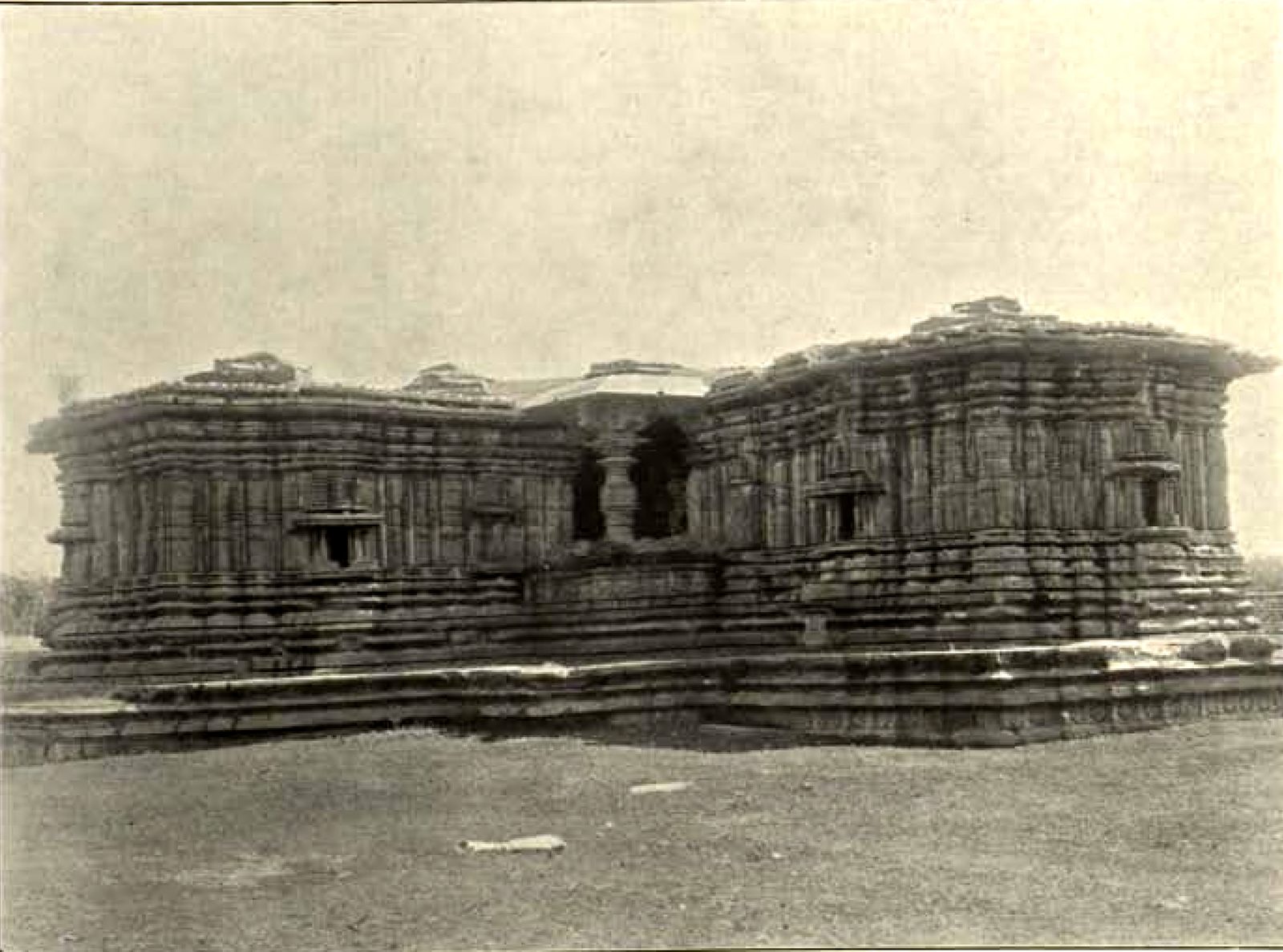 The long, low stone temple with its shrines and a central porch, seen across an open, grassy yard. Deep horizontal mouldings run along the walls.