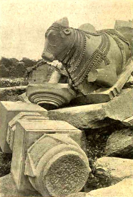 A carved stone bull with a decorated harness lies tilted among fallen pillar parts and broken slabs.