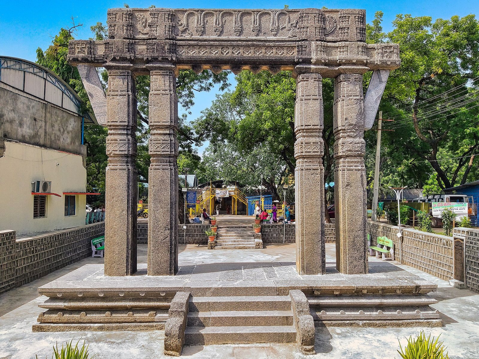 A carved stone gateway of four square pillars and a heavy beam stands on a new stepped stone platform, with trees and buildings behind it.
