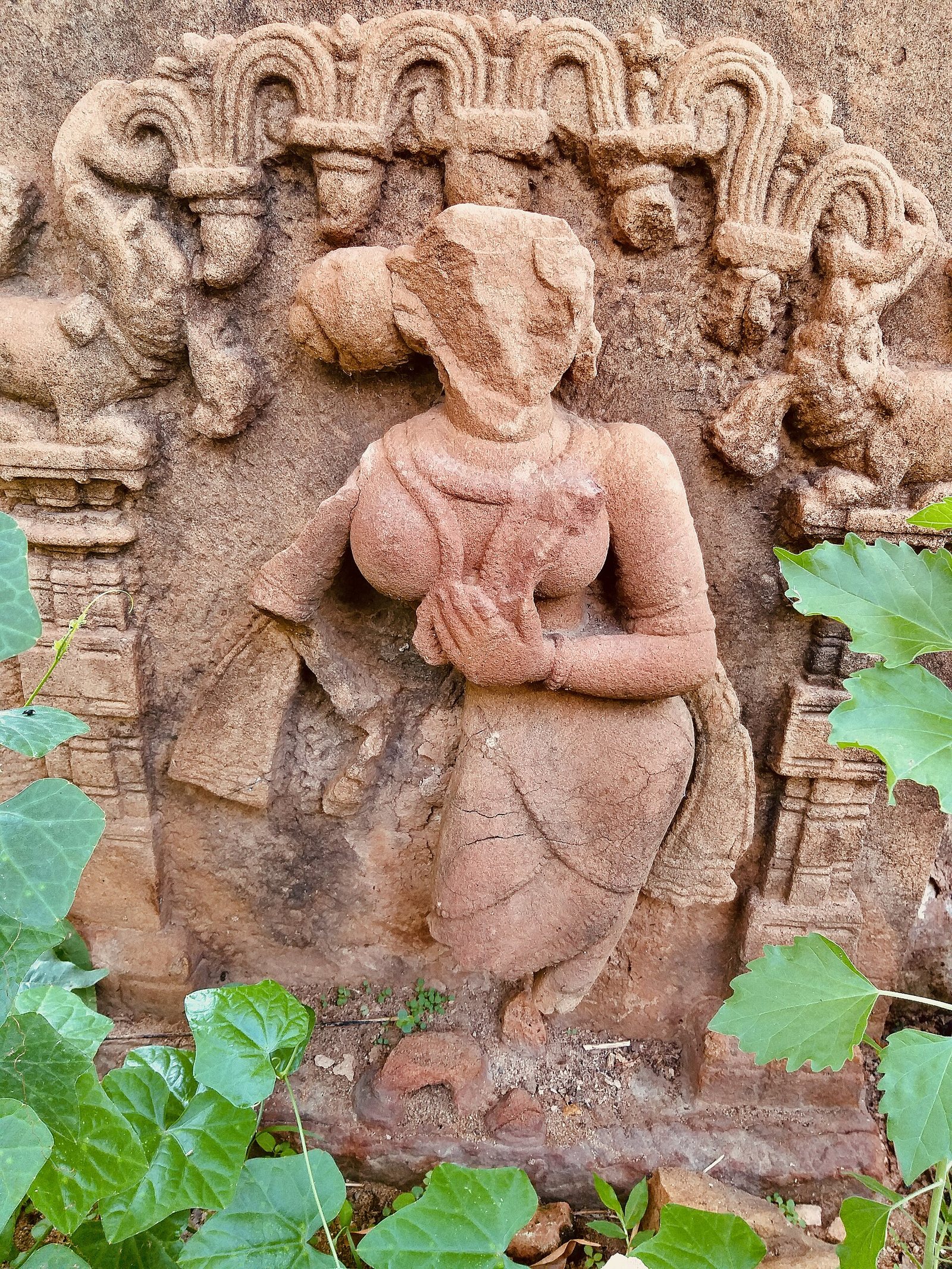 A weathered red stone panel carved with a standing female sculpture under an arch of curling foliage.