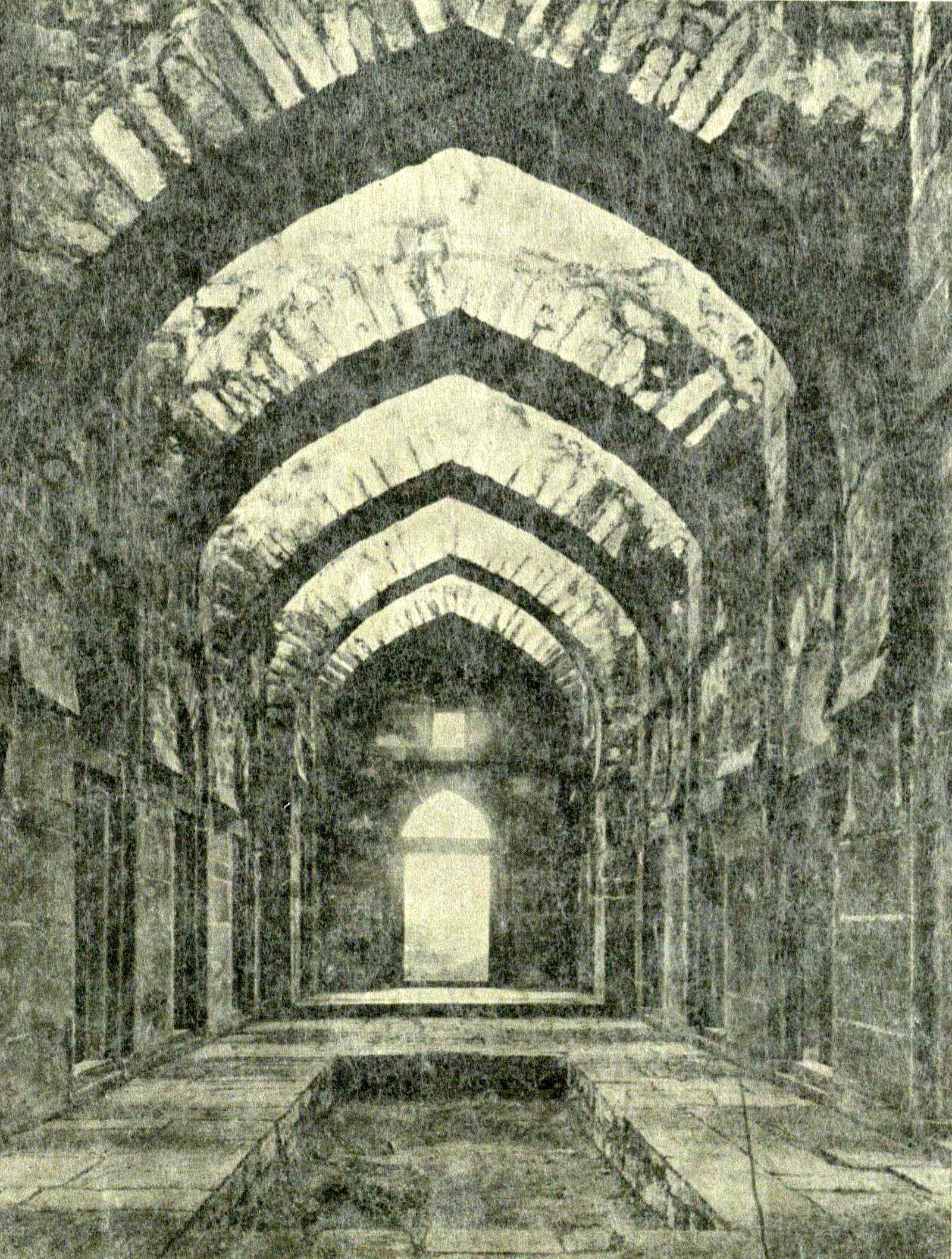 The interior of a long stone hall spanned by a row of pointed stone arches. A sunken channel runs along the middle of the floor. A doorway at the far end lets in light.