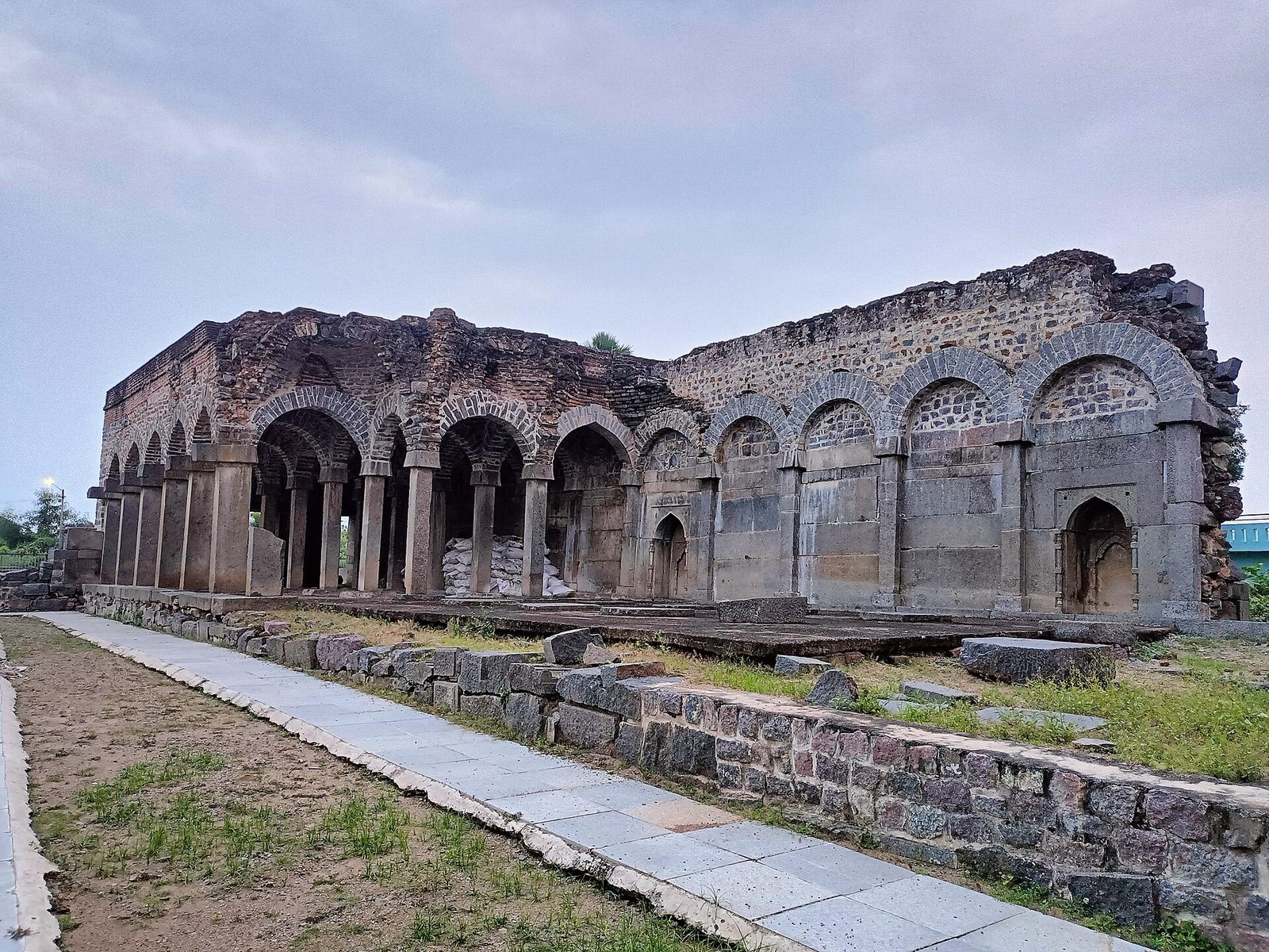 The partly ruined Konda Masjid: a long arcade of pointed arches on stone piers, with collapsed upper walls, behind a paved path and a low rubble wall