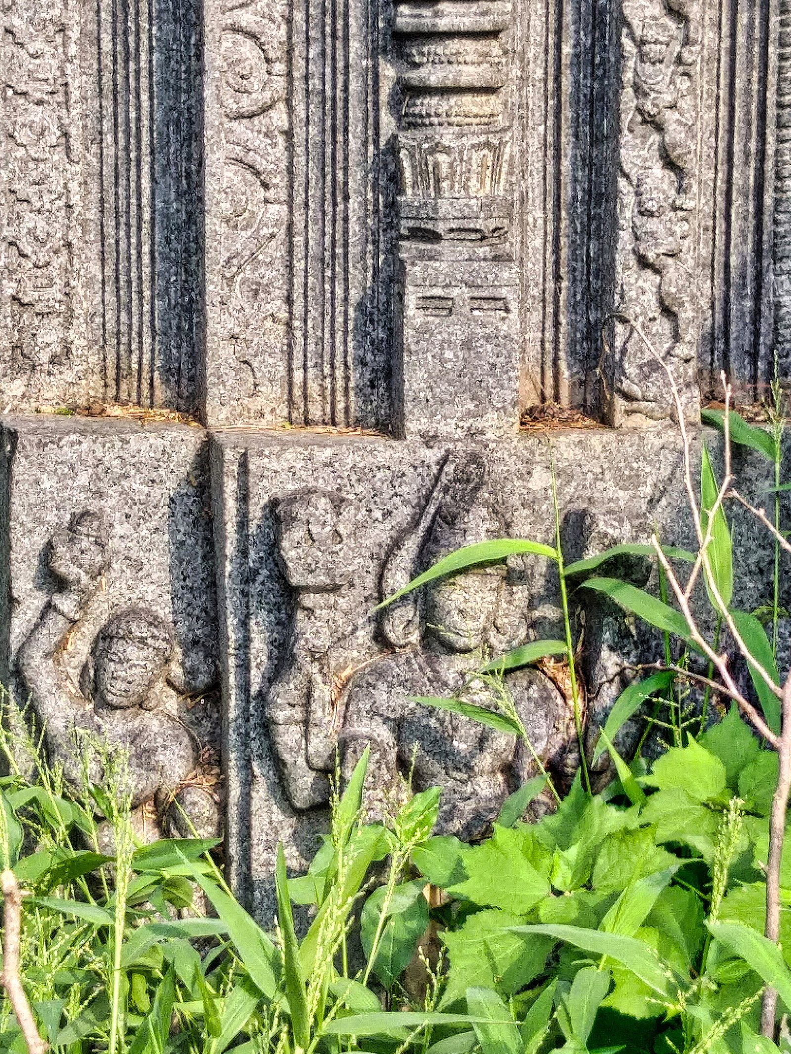 A grey stone pilaster with fluted bands and scrolls. Below it, small reliefs show people in a row, partly hidden by plants.