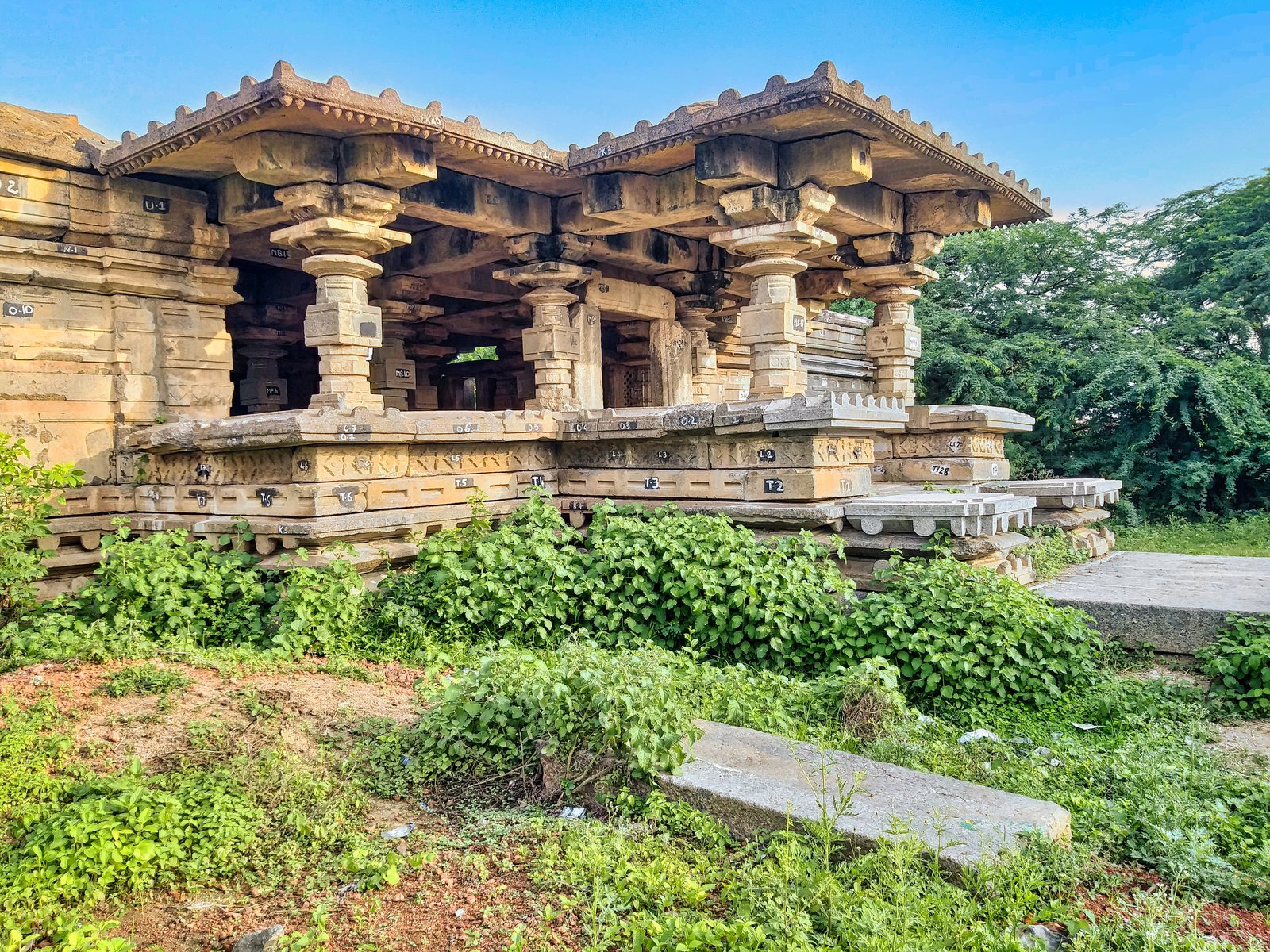 A low stone temple with lathe-turned pillars and heavy beams stands on a moulded platform, with green bushes in front and trees behind.