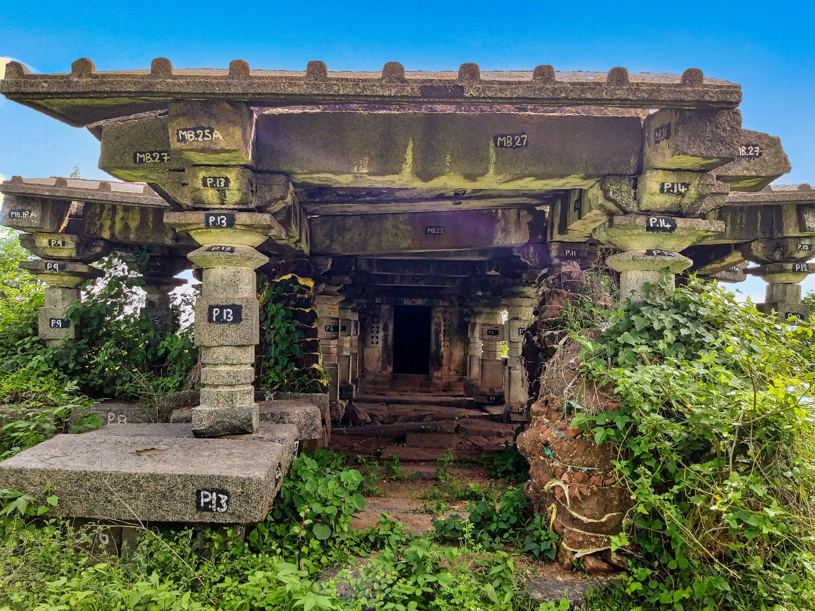 A stone porch on square pillars leads into a dark pillared hall. Plants and an earth mound crowd the entrance, and numbers are painted on the stones.