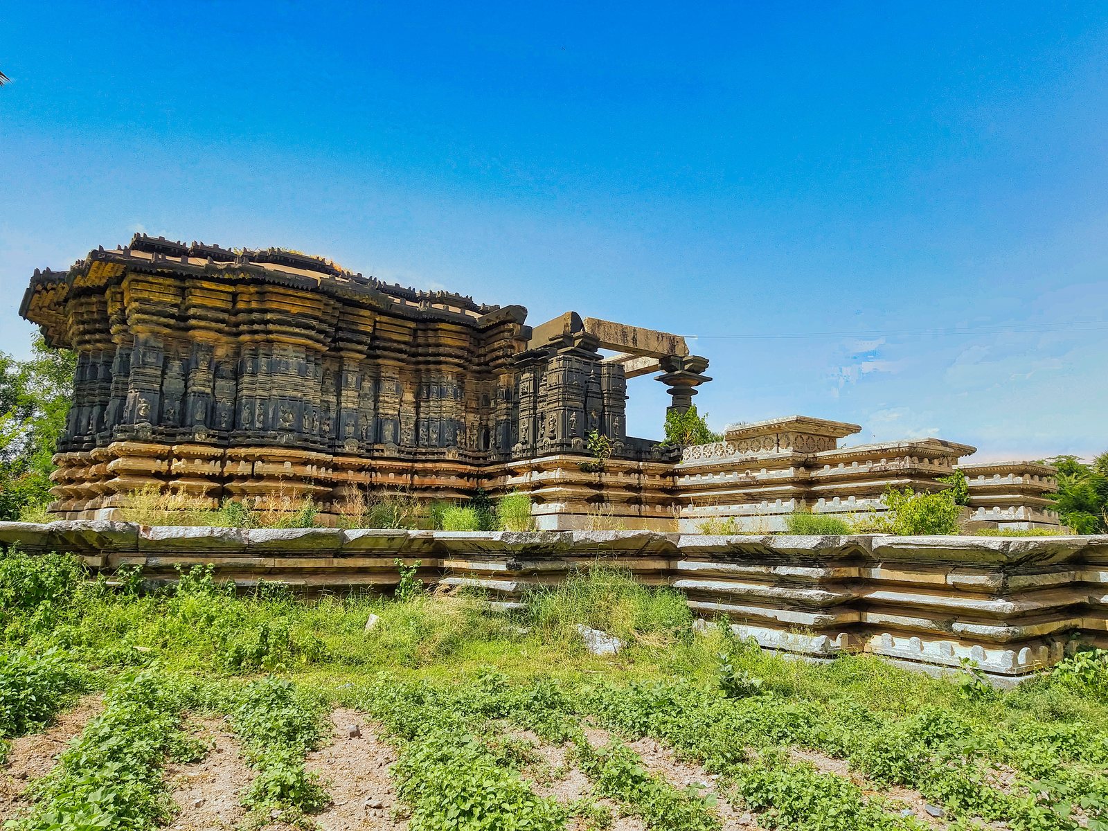 A ruined stone temple with deeply carved dark outer walls stands on a wide, stepped stone platform. Weeds and grass cover the ground in front.