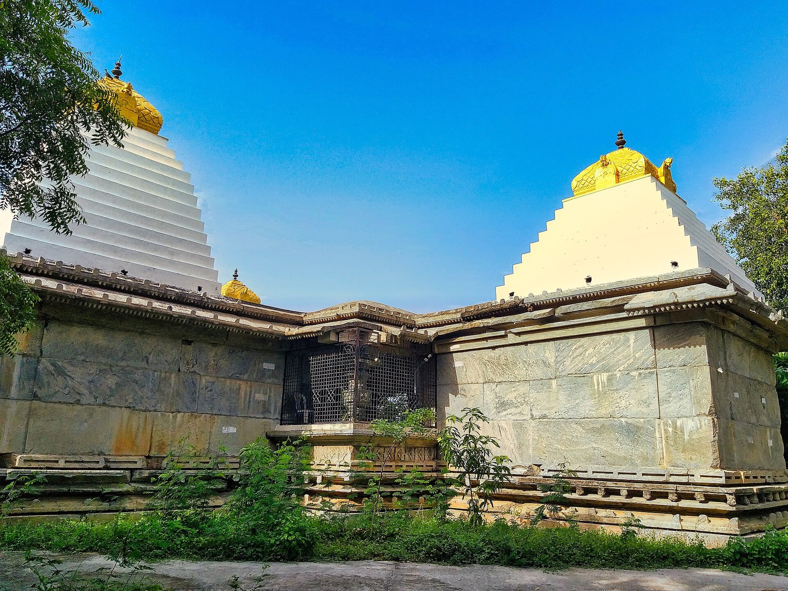 Two shrines with plain stone walls meet at a corner. White, stepped plaster towers with gold-coloured tops rise above them against a blue sky.