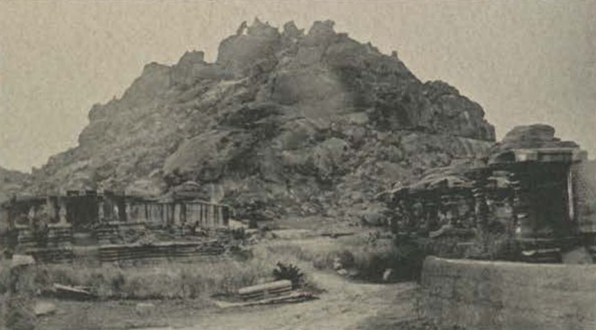 A large rocky hill rises behind two ruined temples, one on the left and one on the right, with a dirt path between them.