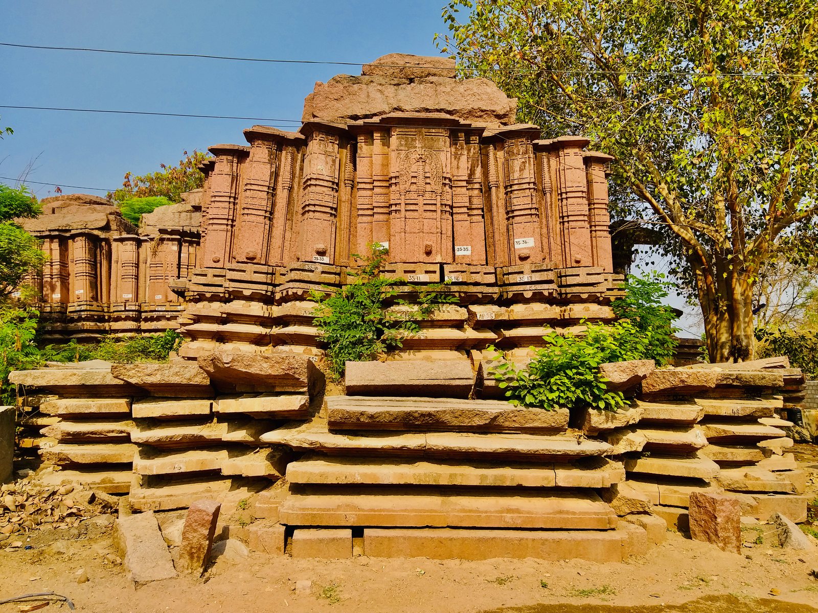 Red stone shrine walls with many projecting pilasters stand on a stepped stone platform. Flat blocks of stone lie on top, and small number labels mark the stones.