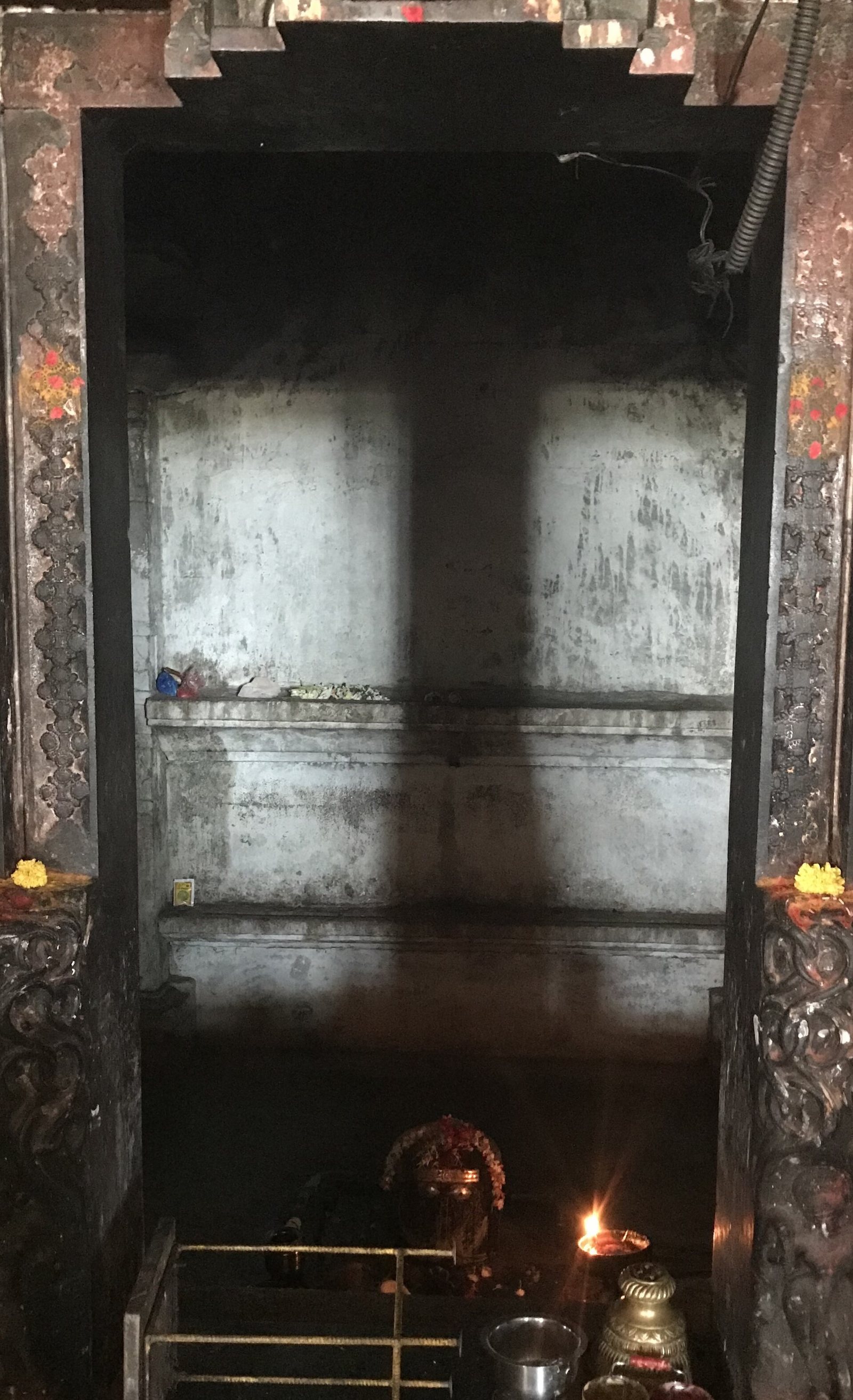 Through a dark doorway, a broad dark band of shadow falls down the pale back wall of the sanctum. A small linga with flowers and an oil lamp stand at the bottom.