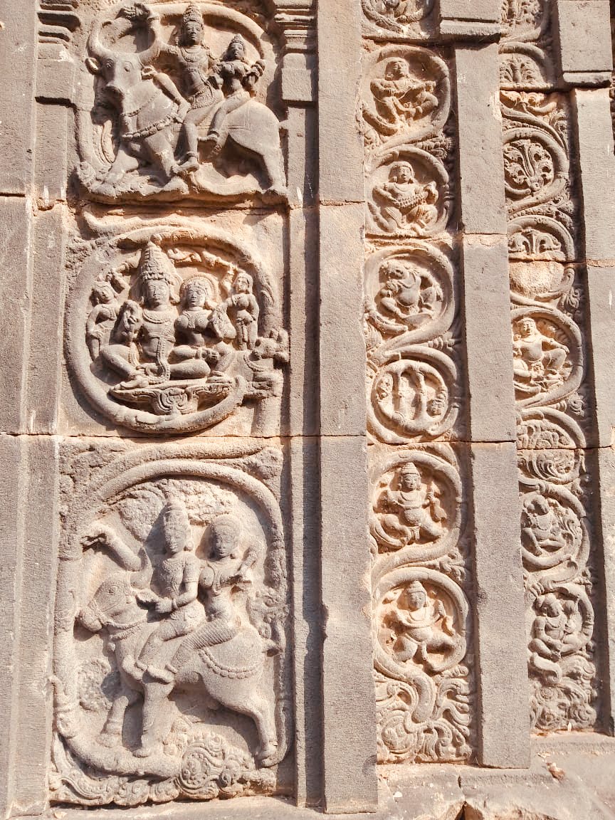A stone wall with tall vertical bands of round scroll medallions, each holding a small carved sculpture. Panels on the left show pairs of deities riding animals.