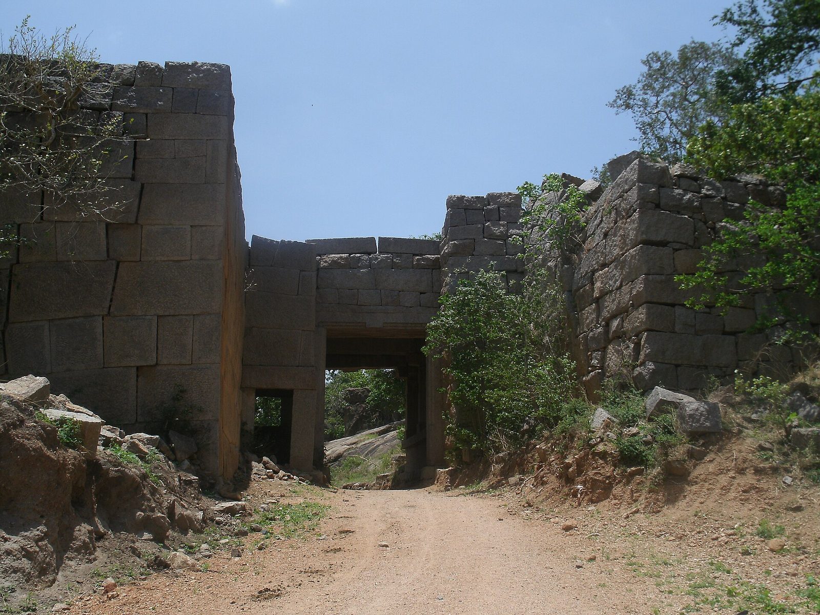 A dirt road runs through a square stone gateway with a flat stone lintel. High walls of large cut granite blocks rise on both sides of the gateway. Shrubs grow over the wall on the right.