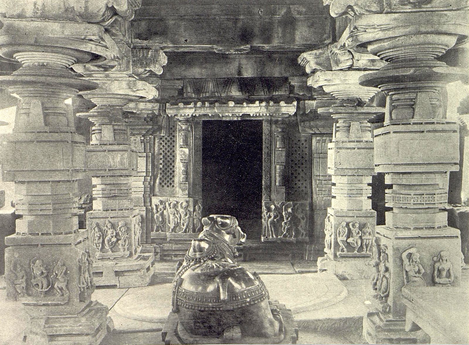 Interior of a stone temple. Heavy carved pillars frame a dark doorway with pierced stone screens on each side. A carved stone bull sits on the floor and faces the doorway.