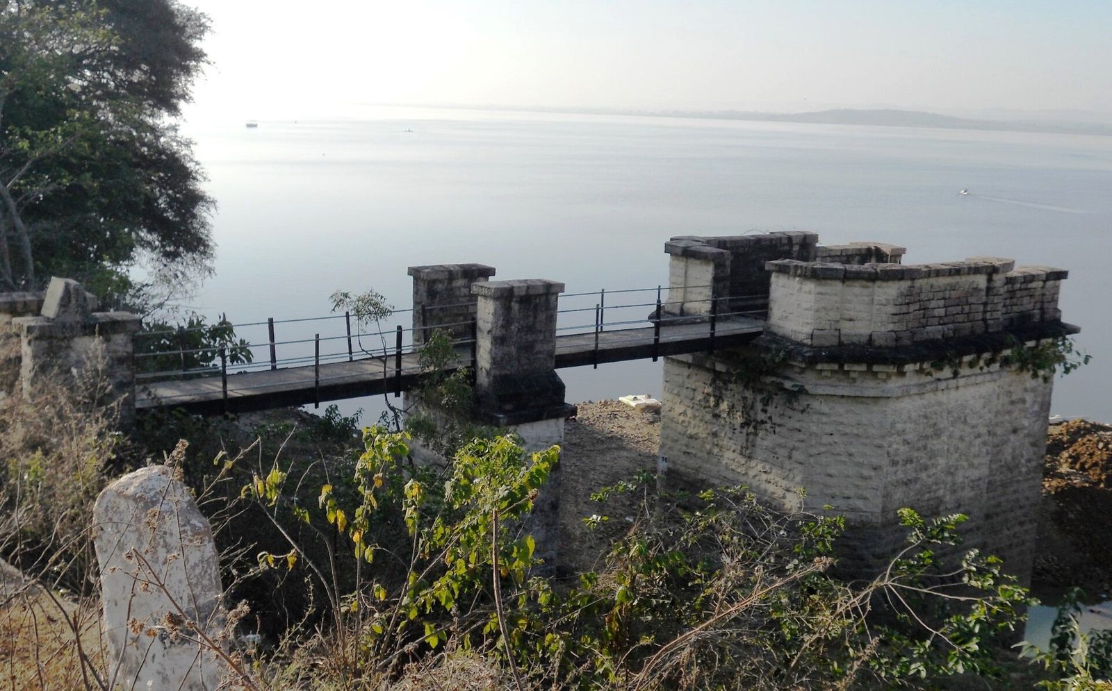 A square masonry tower with a notched parapet stands at the edge of the lake. A narrow metal footbridge on masonry piers links it to the bank. Open water stretches to the horizon behind it.