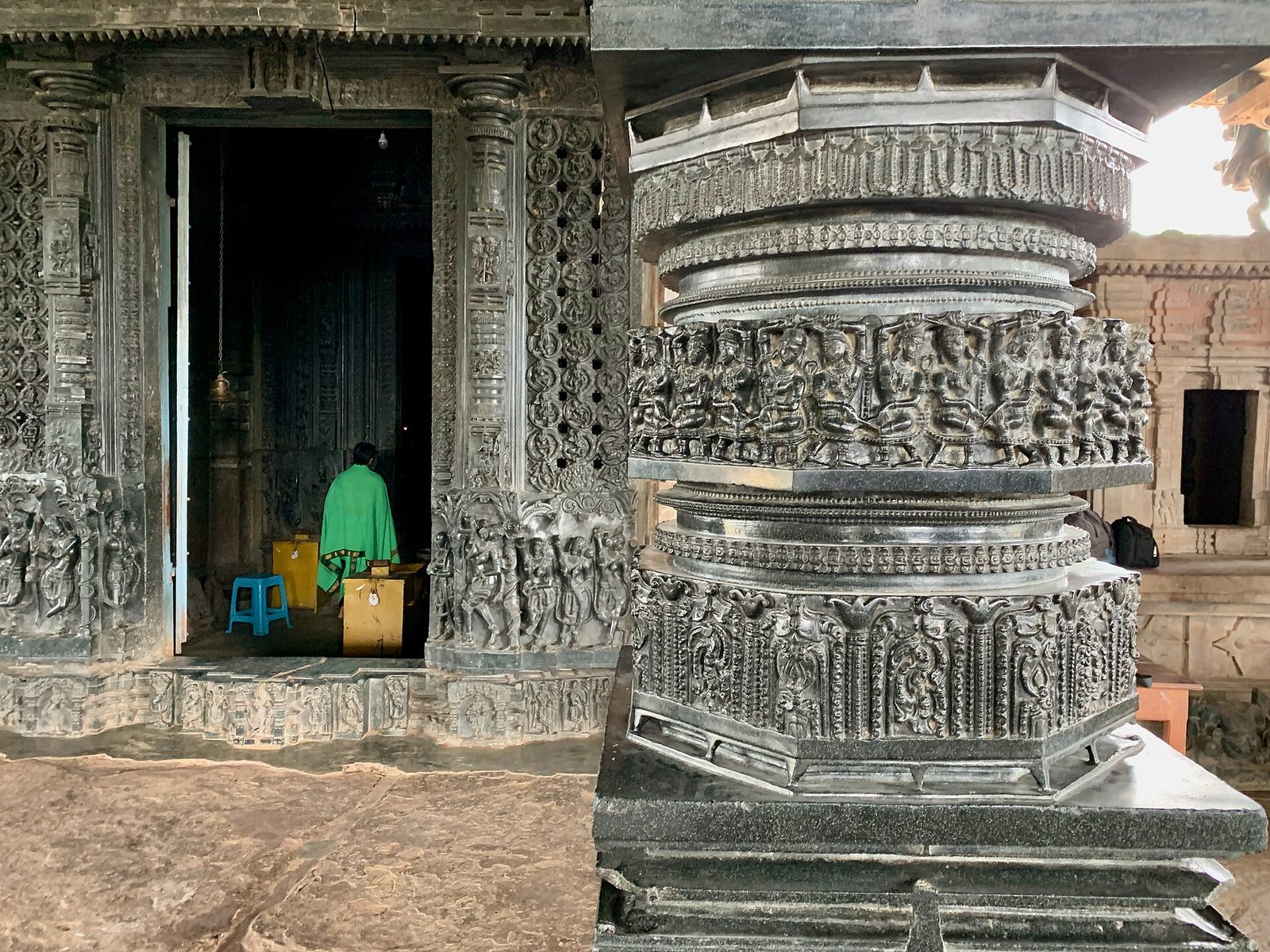 A polished black stone pillar inside Ramappa Temple, lathe-turned into rings and carved with bands of small sculptures, beside a shrine doorway with carved dark stone jambs