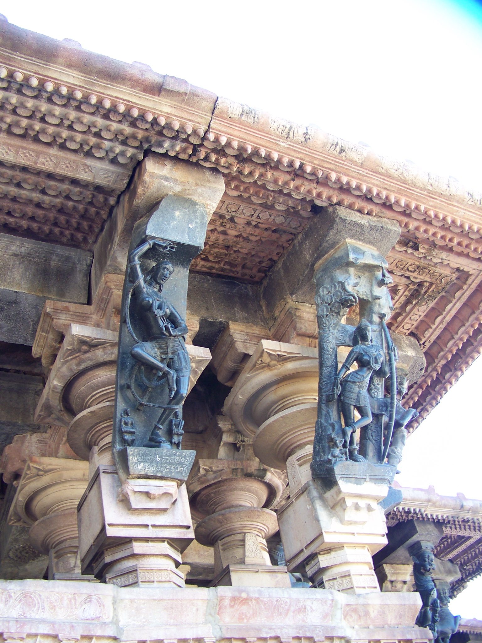Two dark polished stone bracket sculptures of women, set between the column capitals and the overhanging eave of the Ramappa mandapa, seen from below. The sculpture on the right holds a bow.