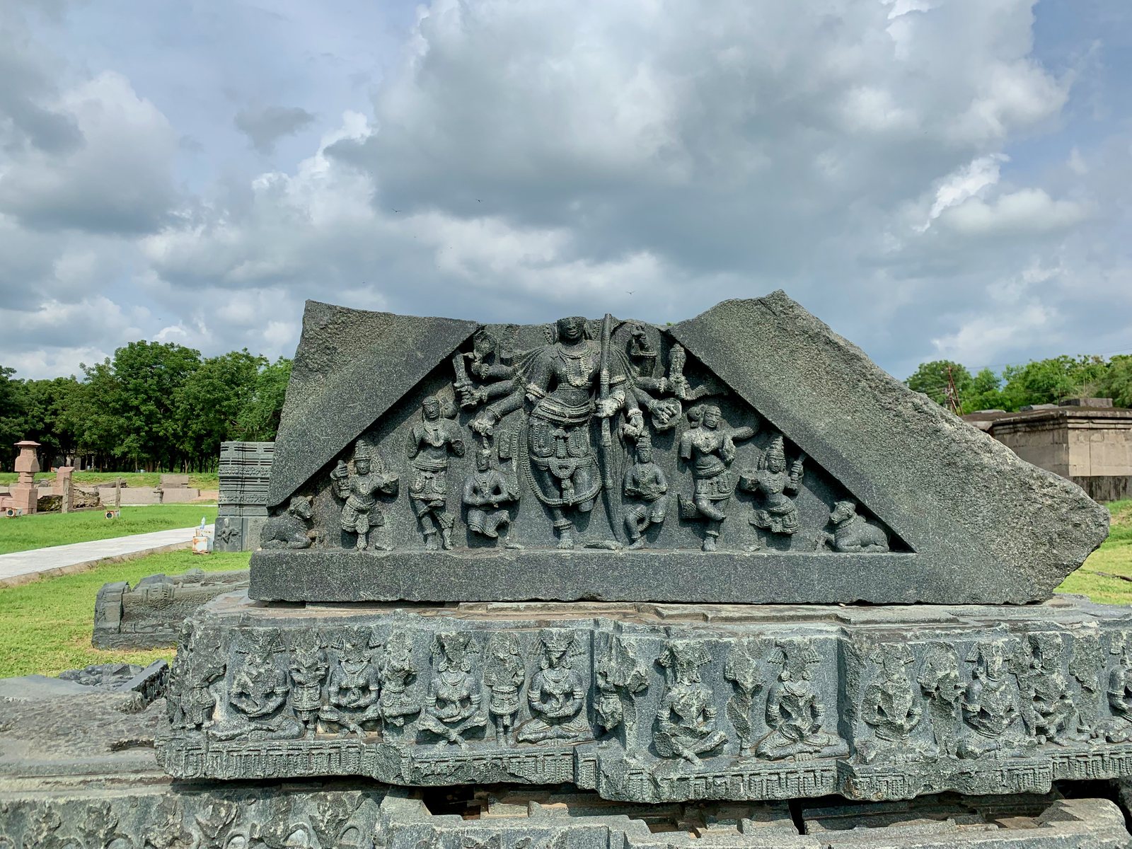 A triangular black stone slab carved with a many-armed dancing god among smaller statues, resting on a carved base on a lawn.
