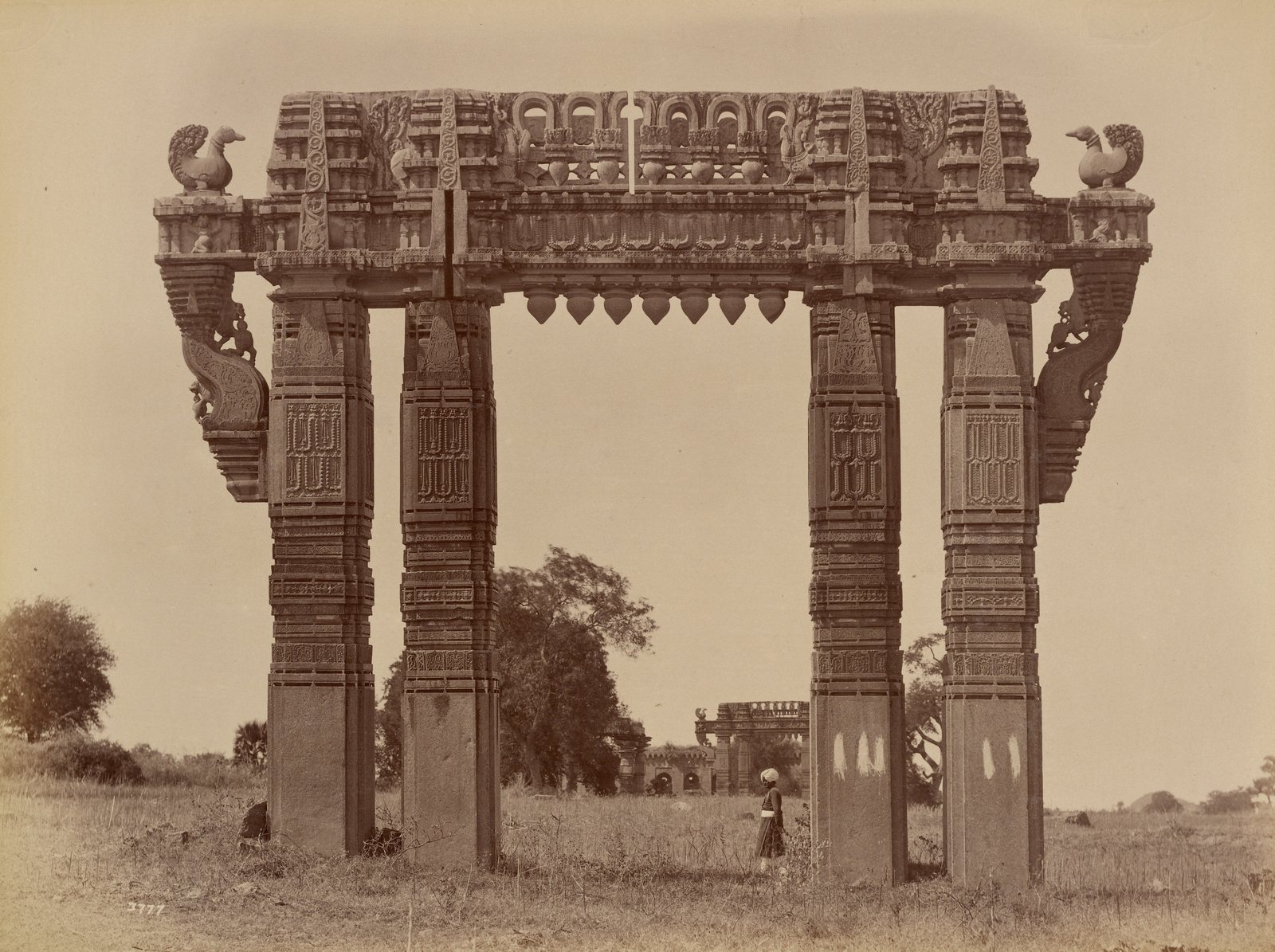 A tall stone gateway of four carved pillars carries a heavy carved beam with a stone swan at each upper corner. A man in a turban stands in the grass under the opening. A second gateway and a long building with arches show in the distance.