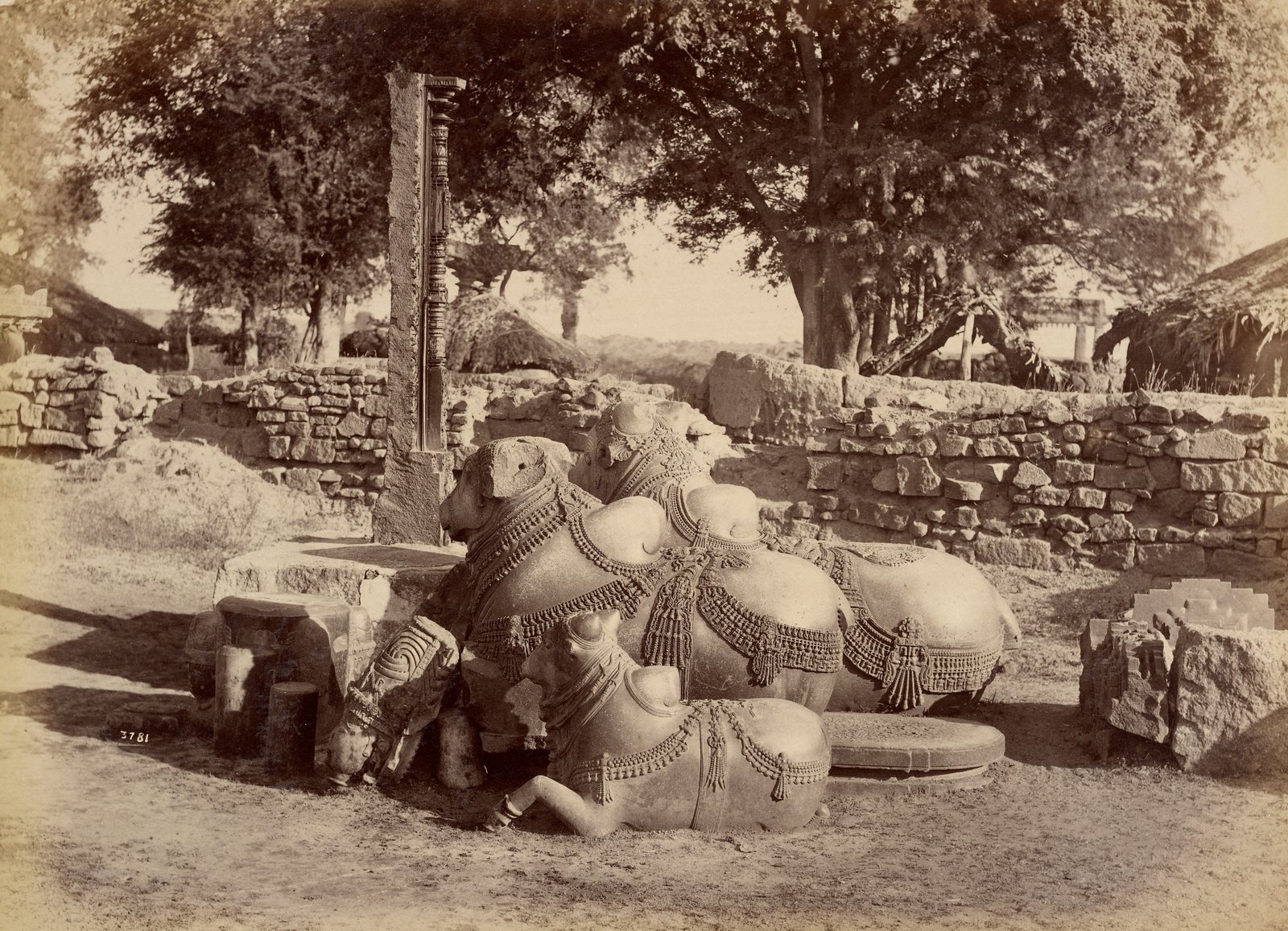 Two large carved stone bulls and a smaller one lie together on bare ground. Carved chains, bells and tassels hang on their bodies. A low wall of rough stones stands behind them, with a tall carved stone post, trees and thatched huts.