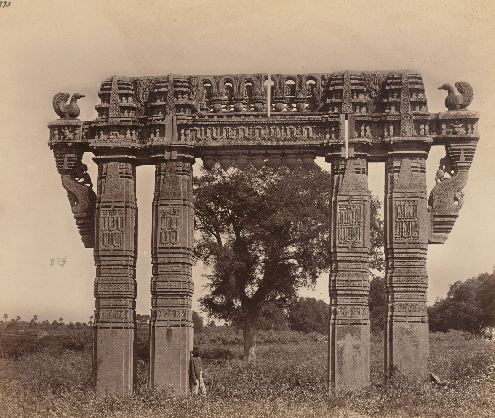 A tall stone gateway of four carved pillars and a carved beam stands in a field of low plants. A stone swan sits at each upper corner, and stone buds hang under the beam. A large tree stands behind the opening, and a man stands beside the inner left pillar.
