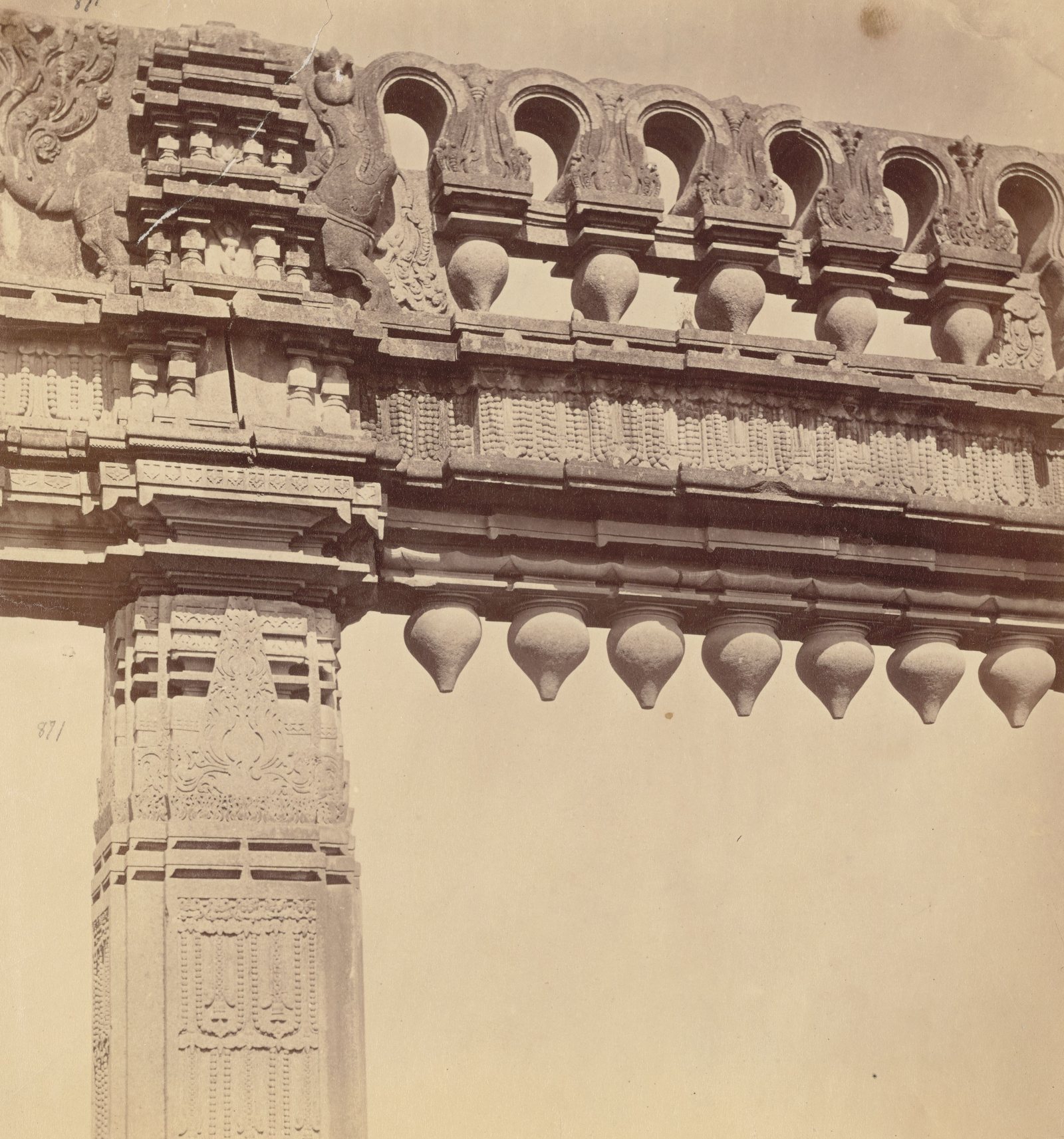 Close view of one end of a carved stone gateway. A row of arched openings with pot shapes runs along the top. A band of beaded carving runs below it, and seven stone buds hang under the beam beside a carved pillar.