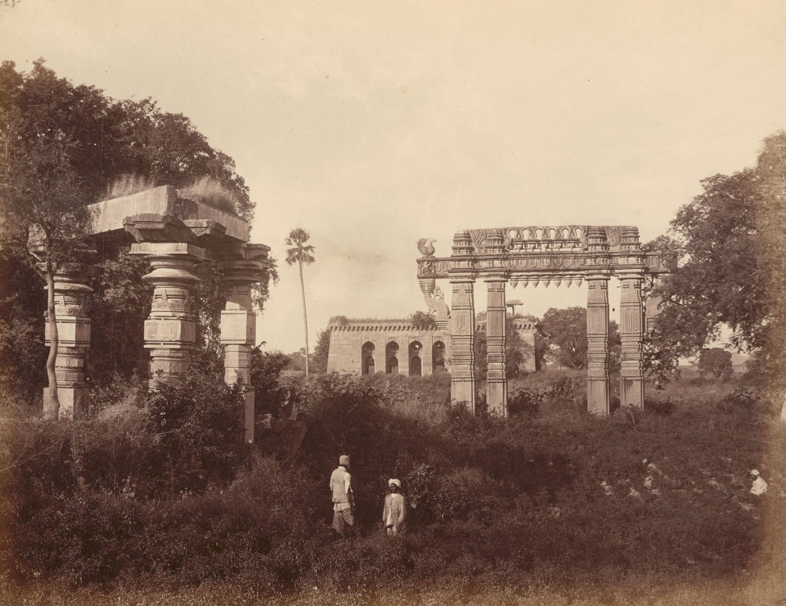 Three carved pillars of a ruined stone hall stand under trees at the left. A carved stone gateway stands in scrub at the right. A long building with arches stands between them in the distance, and three men stand in the scrub in front.