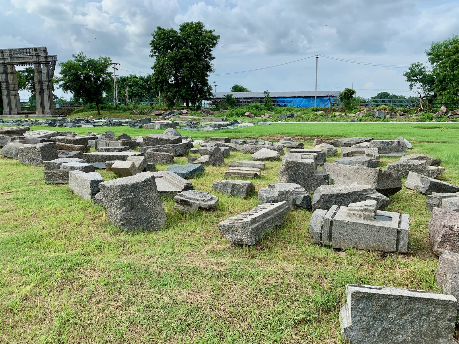 Many dressed and carved stone blocks lie scattered on a green lawn. A stone gateway stands at the far left, and trees and a fence close the view.